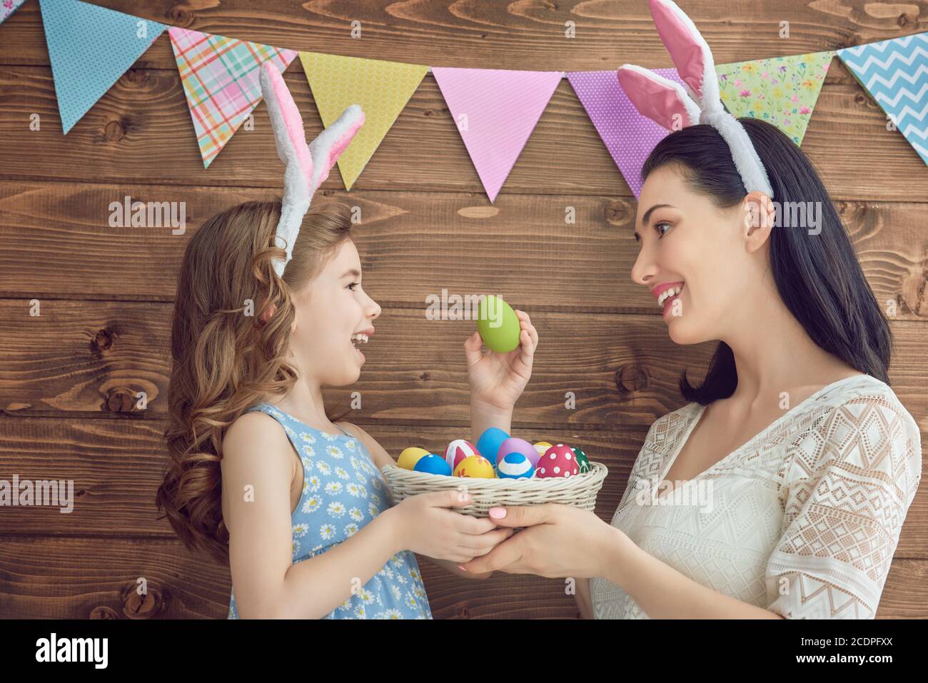 Mother and her daughter with painting eggs. Happy family celebrate Easter. Cute little child girl wearing bunny ears. Stock Photo