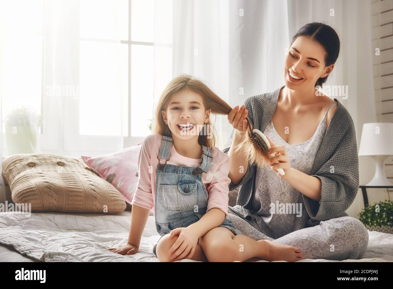 Happy loving family. Mother is combing her daughter's hair sitting on the bed in the room. Stock Photo