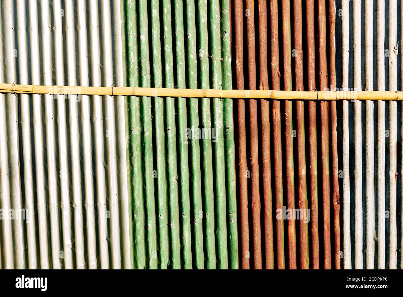 Multicolored painted corrugated wall gives horizontal shades, stripes. One horizontal bamboo stick is holding the rusty fence together. Stock Photo