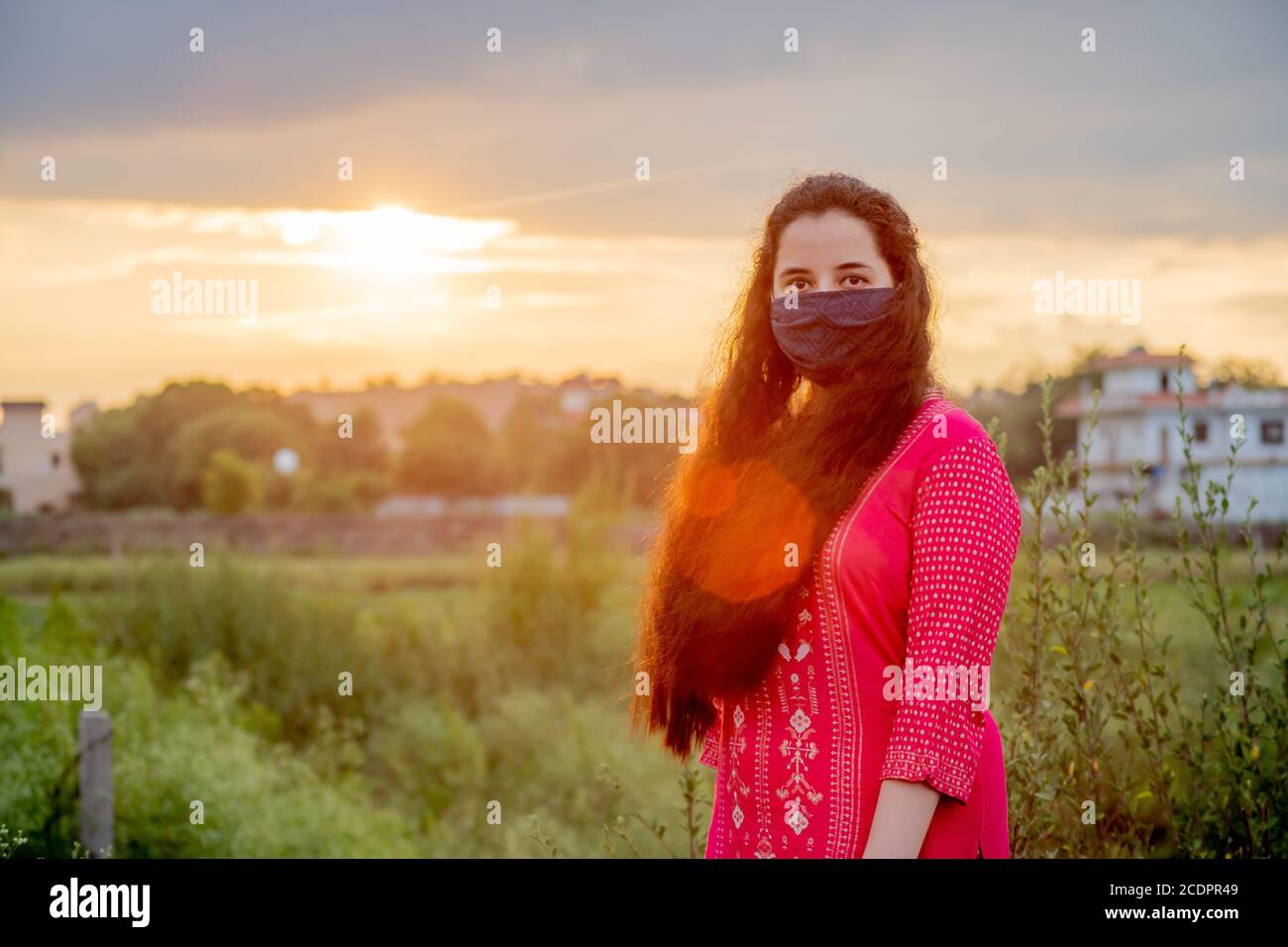 Young Indian girl wearing a mask, indian salwar kameez standing with a field and a beautiful sunset behind her Stock Photo