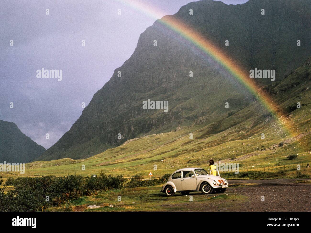 Tourists in a VW Beetle stop in Glencoe, Scotland, to view a nearby rainbow in front of the rockface of Aonach Dubh. Archive image c.1980s. High resolution scan from transparency, August 2020. Credit: Malcolm Park/Alamy. Stock Photo