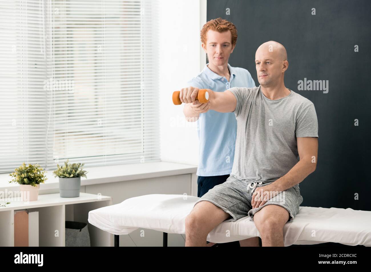Young contemporary physiotherapist helping mature male patient lifting dumbbell Stock Photo