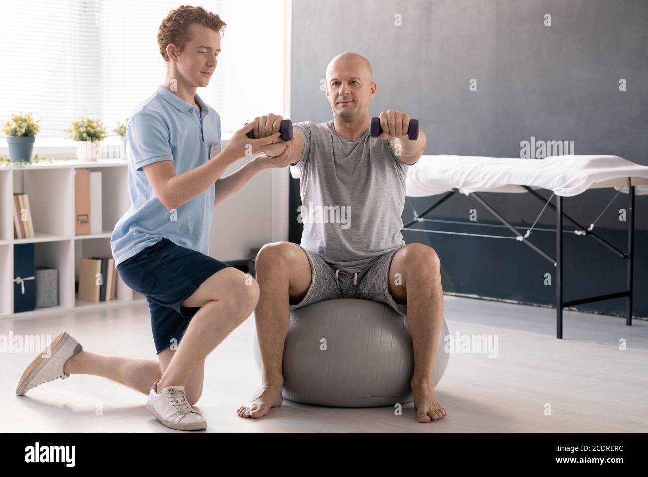 Young physiotherapist in casualwear helping male patient to do exercise Stock Photo