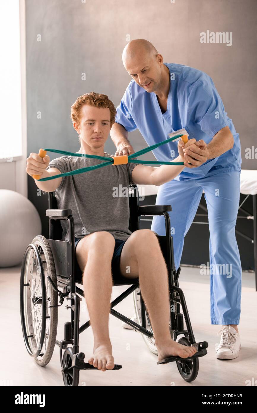 Mature clinician helping young patient in wheelchair while supporting his hand Stock Photo