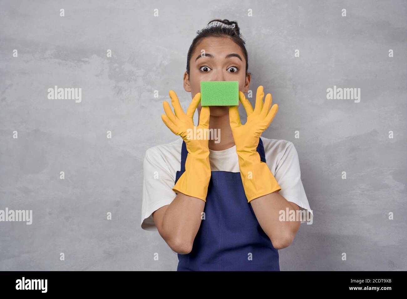 Funny cleaning lady in uniform and rubber gloves playing with kitchen sponge, looking at camera and smiling while standing against grey wall. Studio shot. Housekeeping, cleaning services Stock Photo