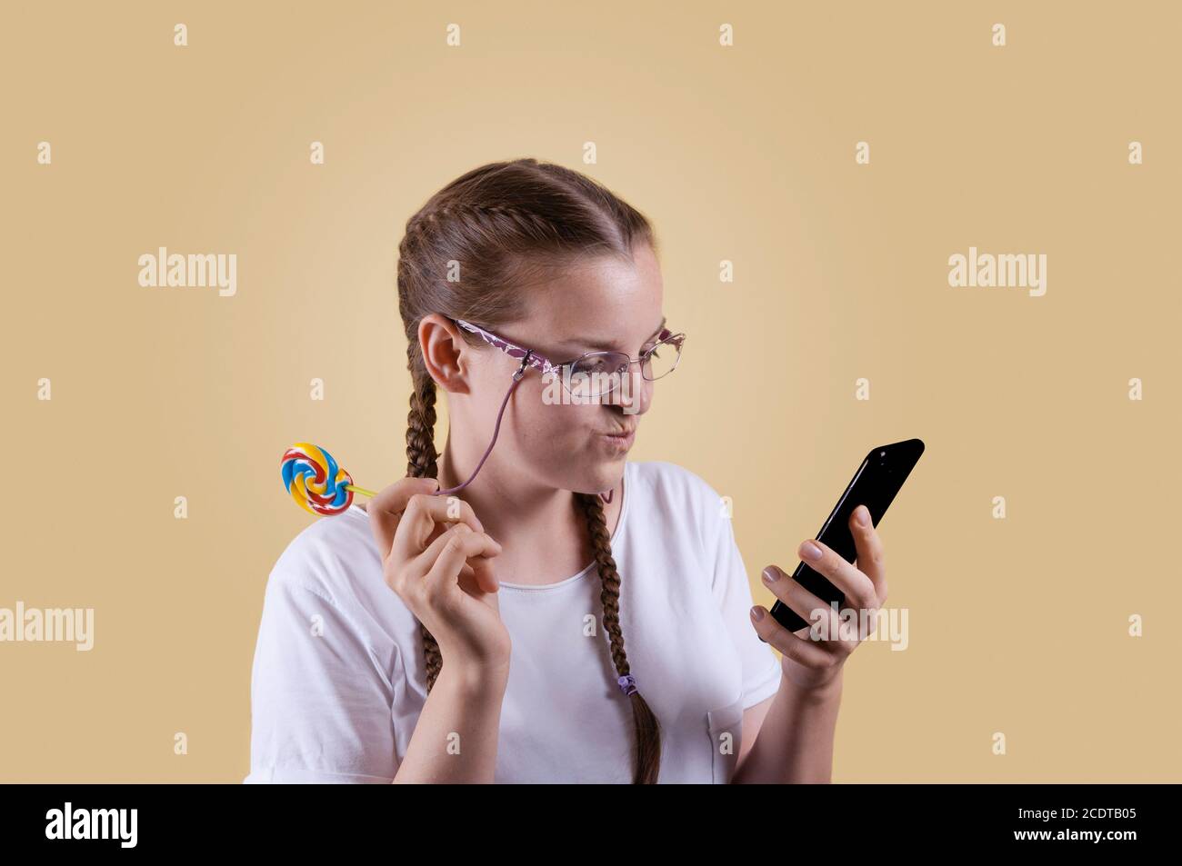 The girl looks at the phone. The girl is holding the phone. Girl with pigtails. The girl is shot against a yellow background. The girl smiles at the n Stock Photo