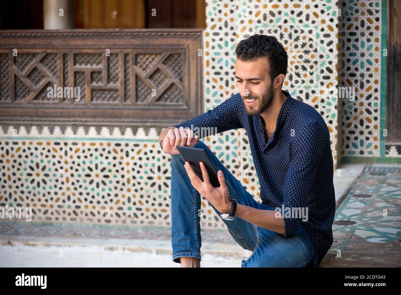 Young Muslim man in casual clothing smiling and working on tablet in traditional Arabian ambient Stock Photo