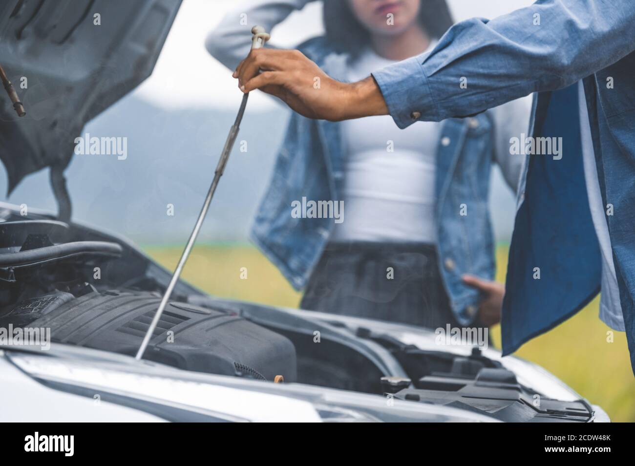 Asian man open car hood for repair as maintenance service with woman background between long road trip. Car broken and breakdown in mountain meadow wi Stock Photo