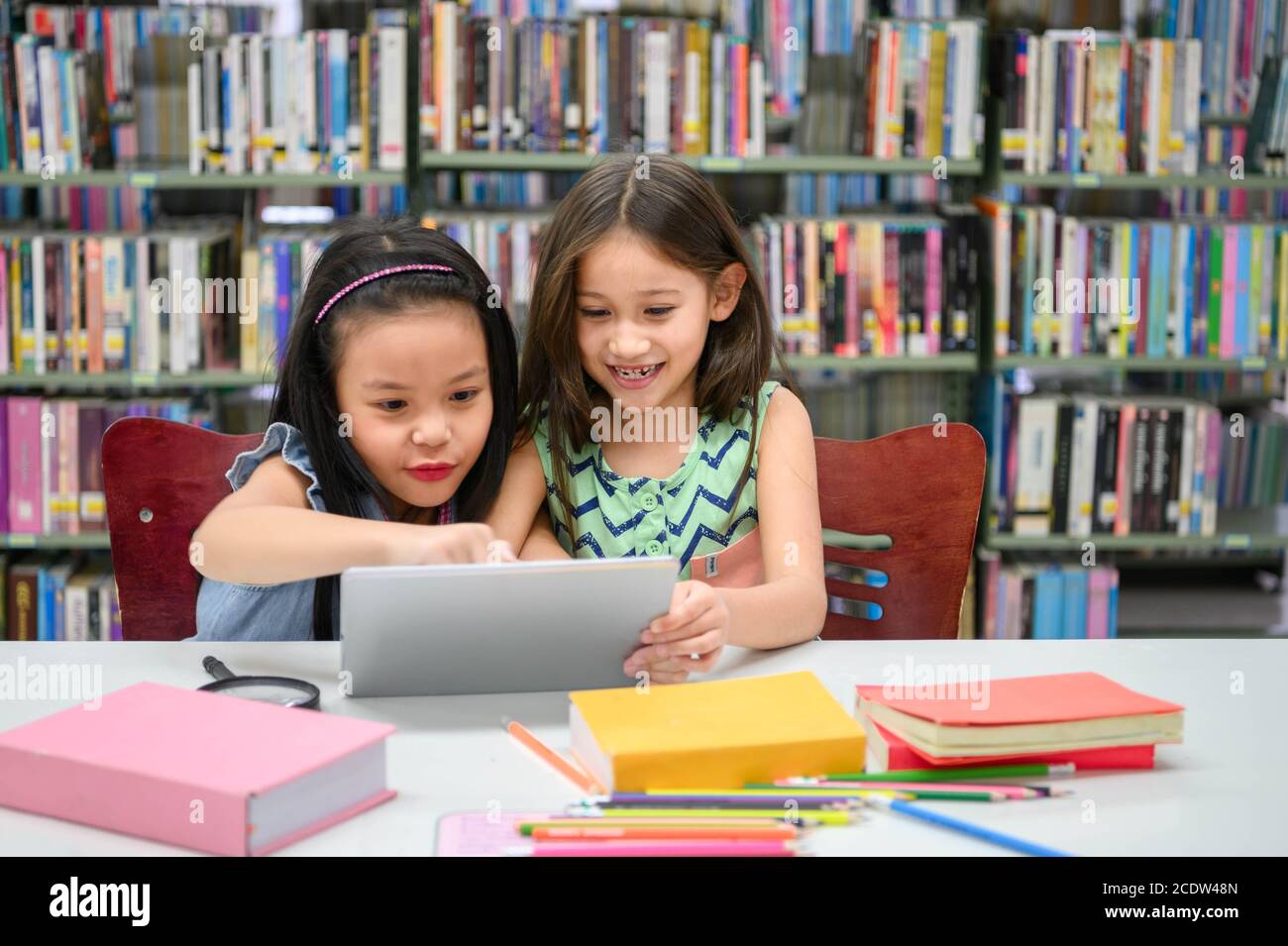 Two little happy cute girls playing on a tablet PC computing device in library at school. Education and self learning wireless technology concept. Peo Stock Photo