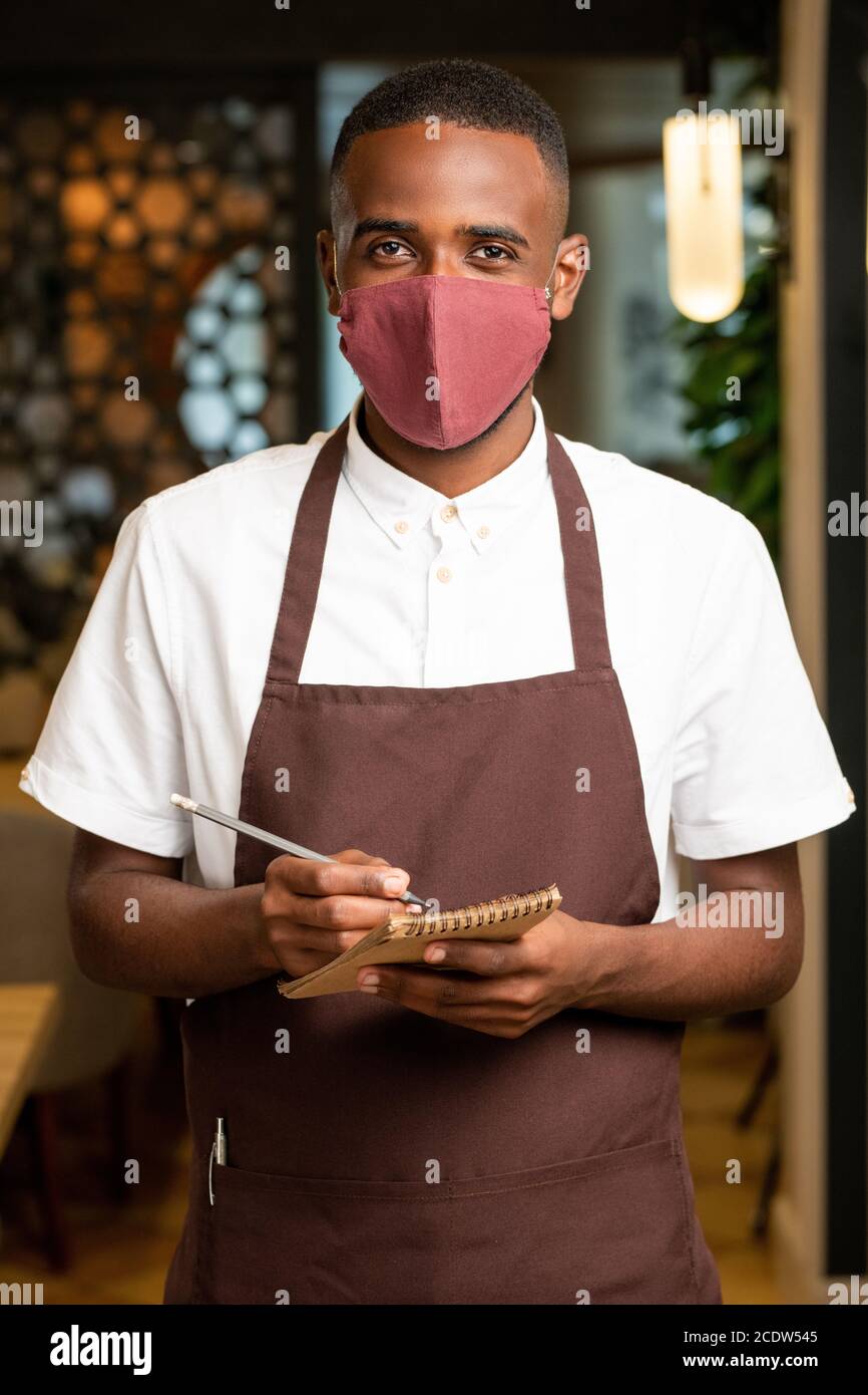 Happy young African man in brown apron and protective mask taking orders Stock Photo