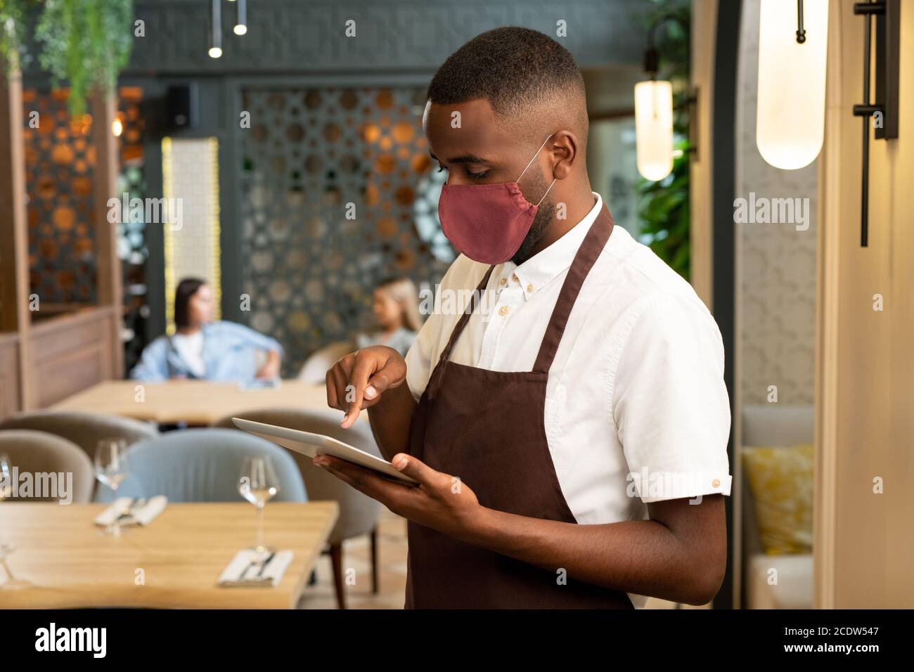 Young African man in brown apron and protective mask taking online orders Stock Photo