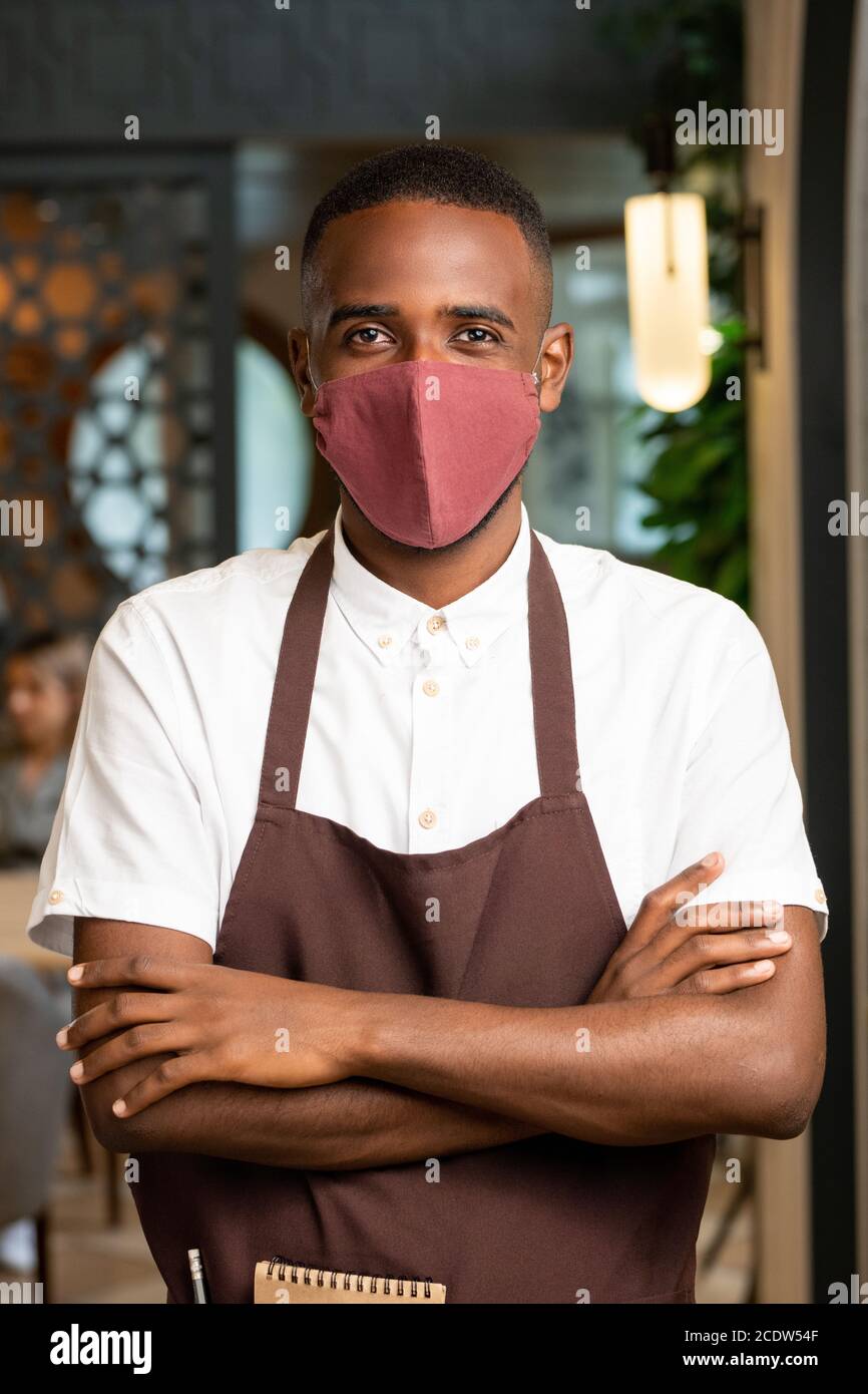 Happy young man of African ethnicity in brown apron and protective mask Stock Photo
