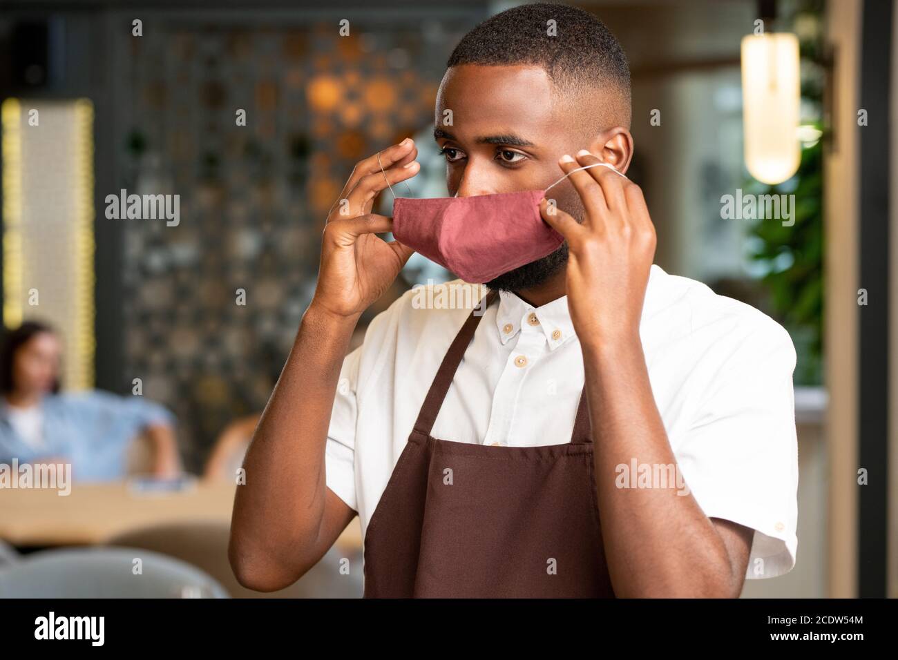 Young African man in uniform of waiter putting protective mask on his ...