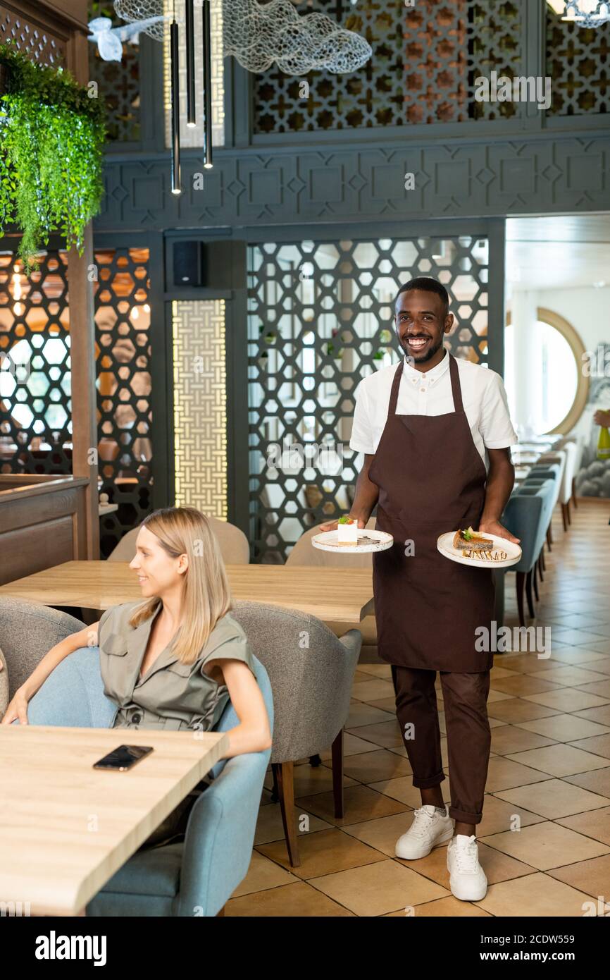 Smiling young waiter of African ethnicity carrying plates with orders of females Stock Photo