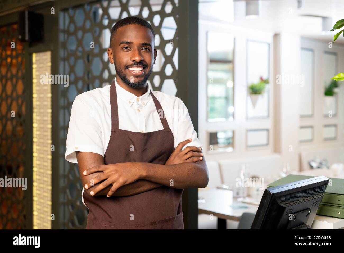Young cheerful waiter of African ethnicity in uniform looking at you with smile Stock Photo