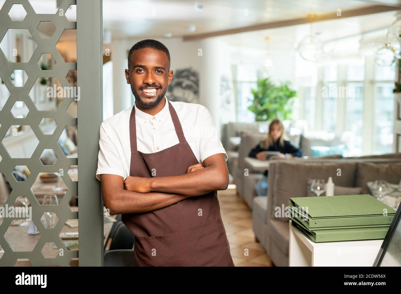 Cheerful young African waiter in workwear leaning against door in restaurant Stock Photo