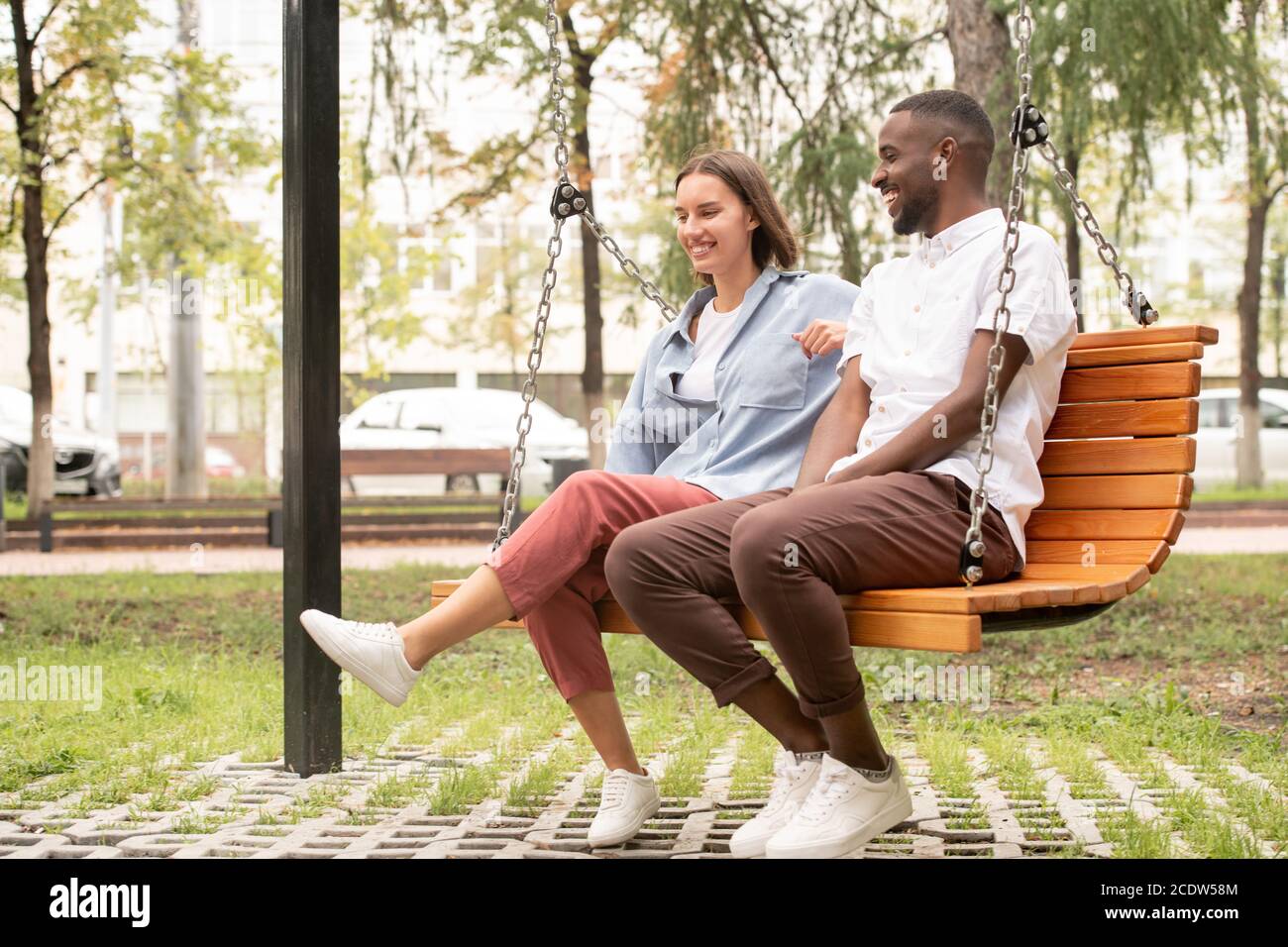 Happy young intercultural couple in casualwear sitting on wooden swing in park Stock Photo