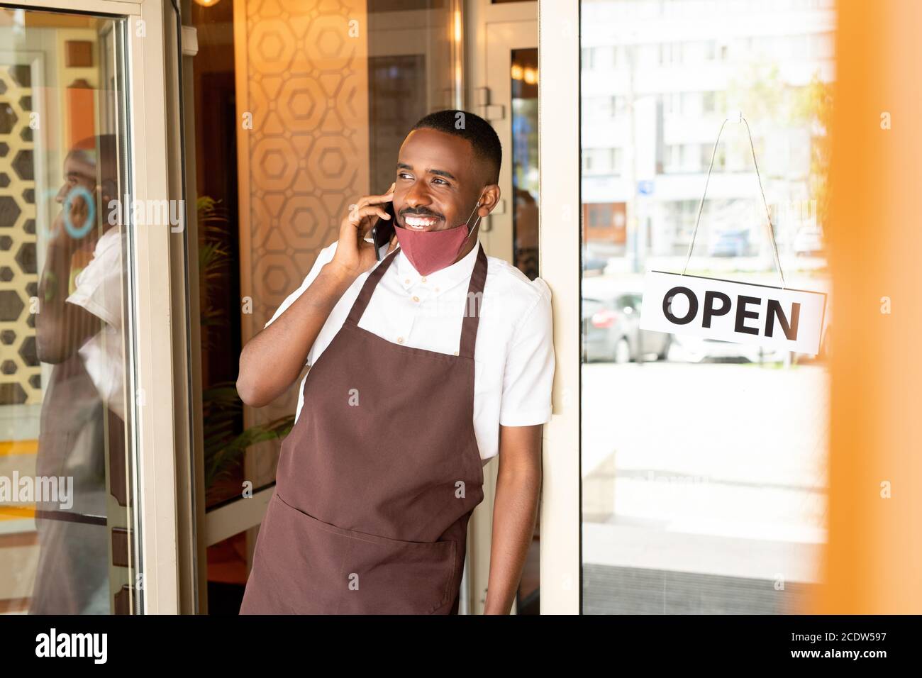 Happy young waiter in uniform and protective mask standing in the door of cafe Stock Photo