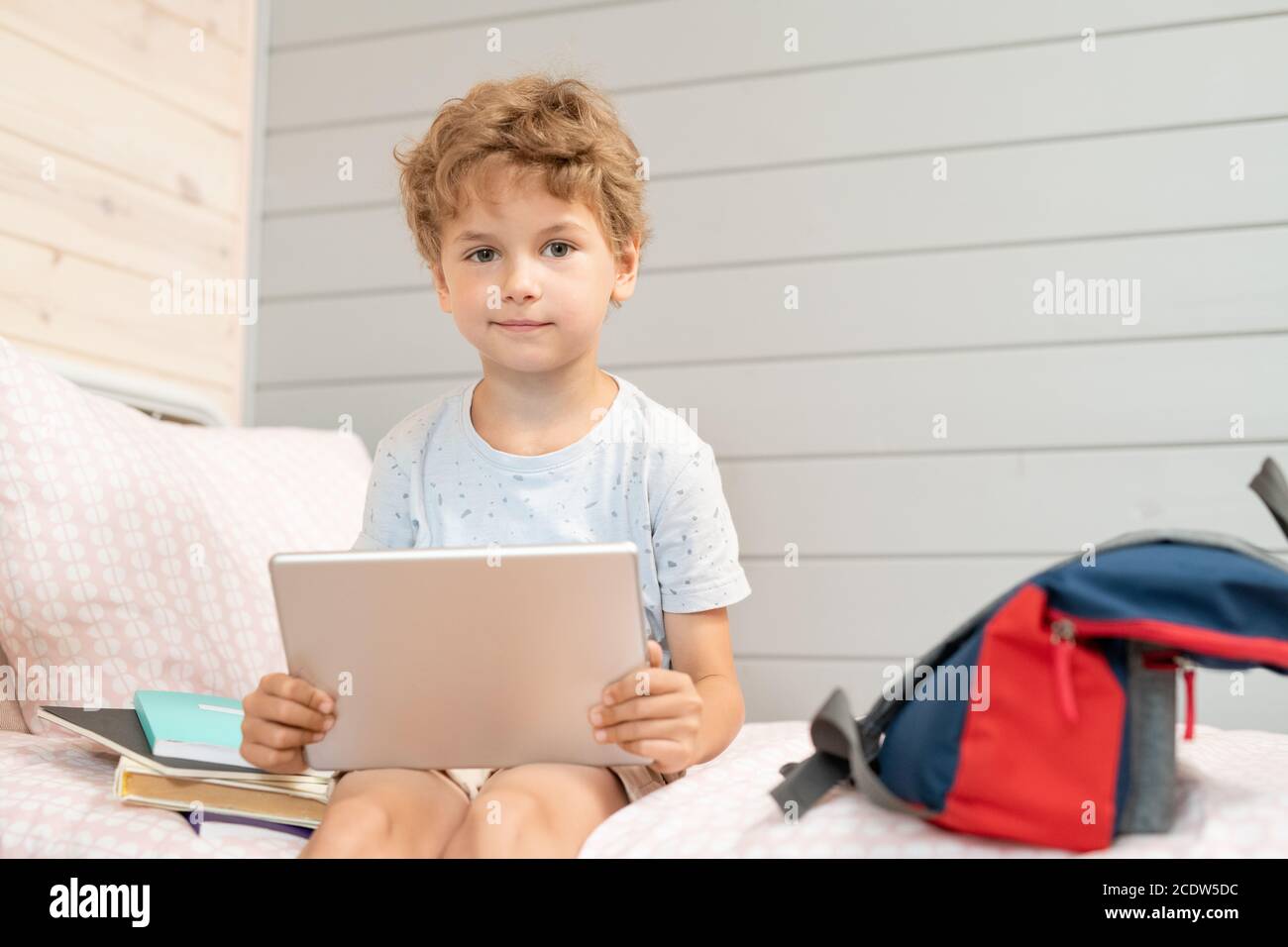 Adorable blond schoolboy with digital tablet sitting on bed after school Stock Photo