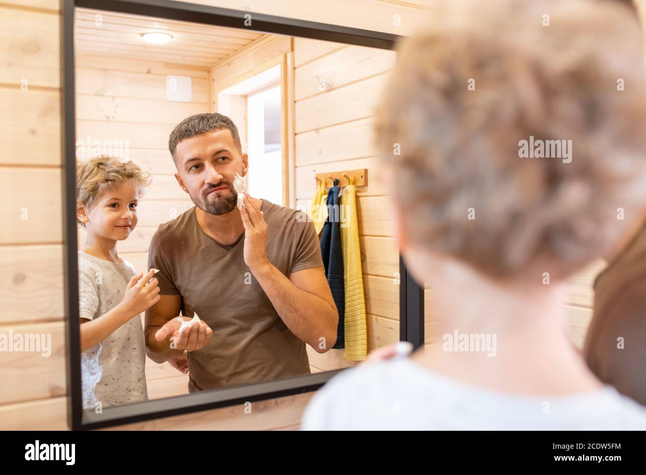 Cute little boy with blond curly hair looking at father applying foam on face Stock Photo