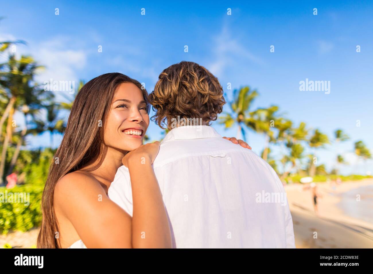 Couple hugging on tropical beach Asian woman holding man looking away pensive. Beautiful girlfriend happy smiling at sunset with boyfriend Stock Photo