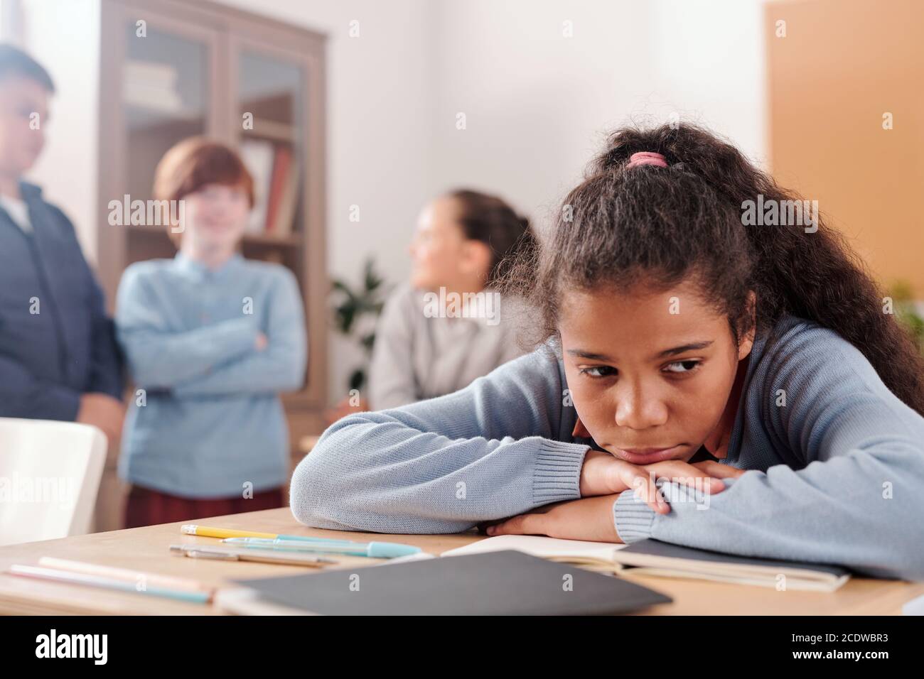 Upset or offended schoolgirl sitting by desk against classmates talking Stock Photo