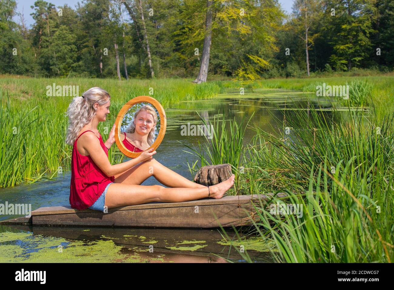 Young woman sits with mirror at water in nature Stock Photo