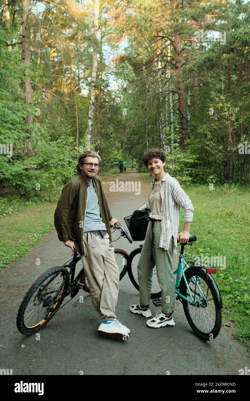 Young joyful couple with bicycles standing between trees in front of camera Stock Photo