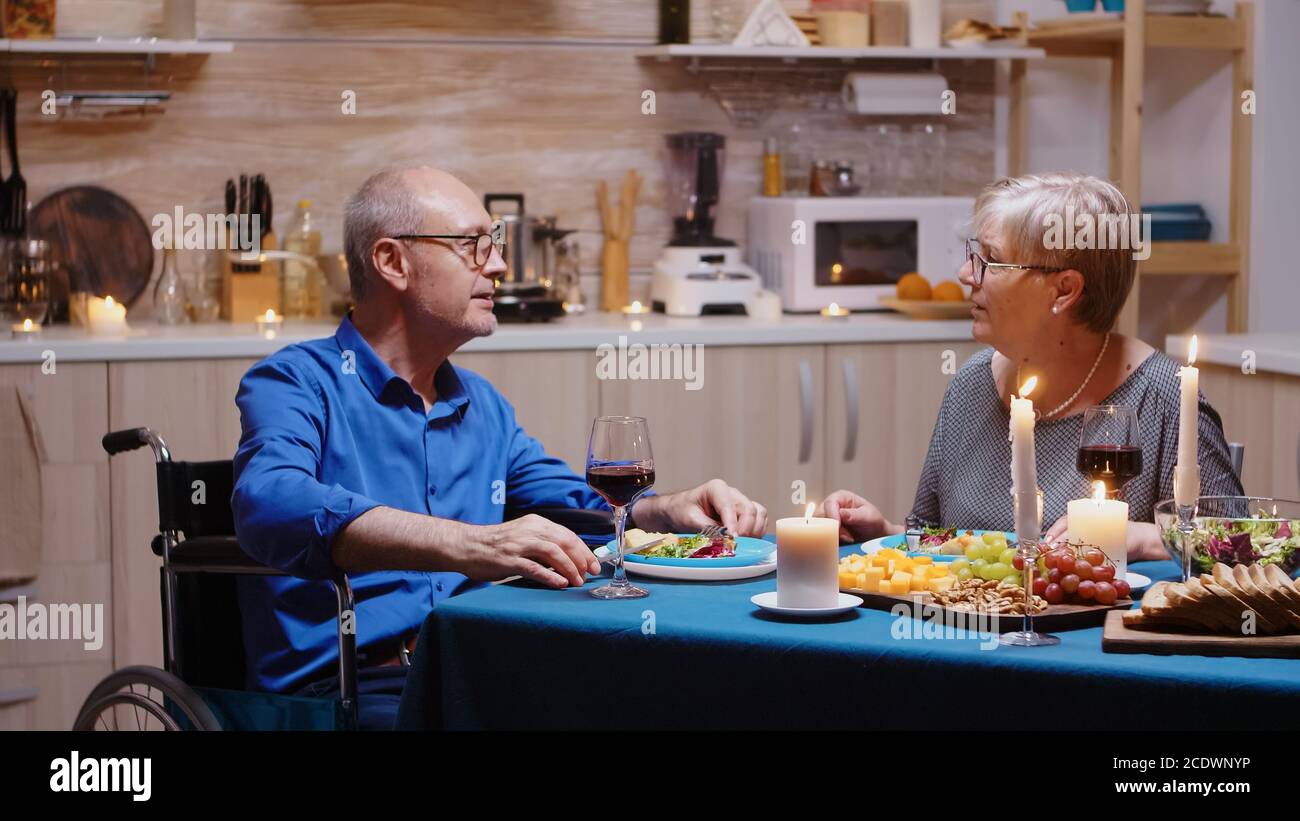 Aged senior man with disabilities having romantic dinner sitting at the table in cozy kitchen. Wheelchair immobilized paralyzed handicapped man dining with wife at home, enjoying the meal Stock Photo