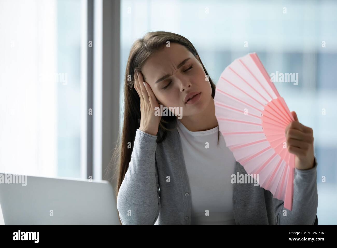 Overheated unhappy young businesswoman waving paper fan in office Stock Photo
