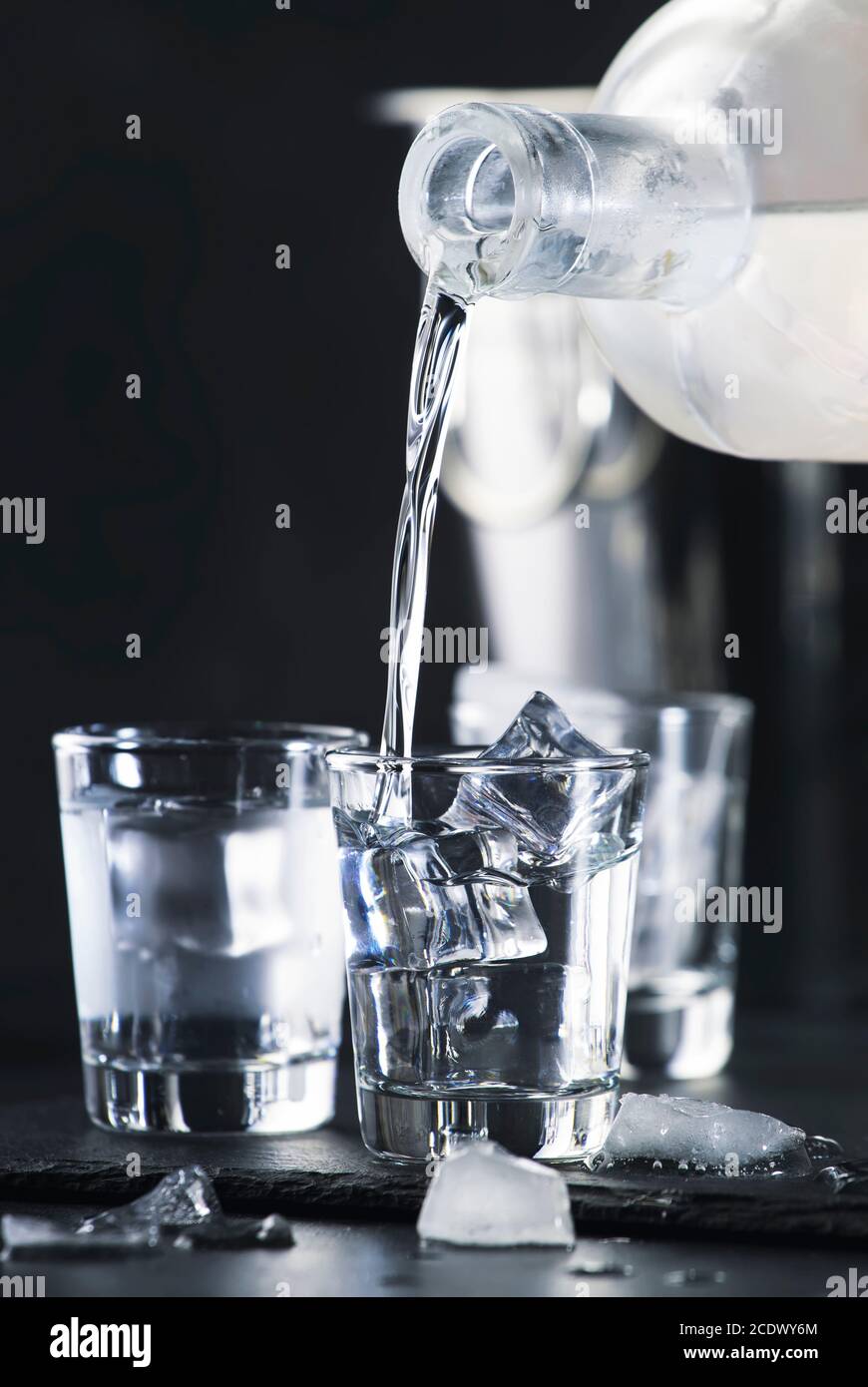 Vodka in shot glasses pouring out of the bottle on black stone background, iced strong drink in misted glass Stock Photo