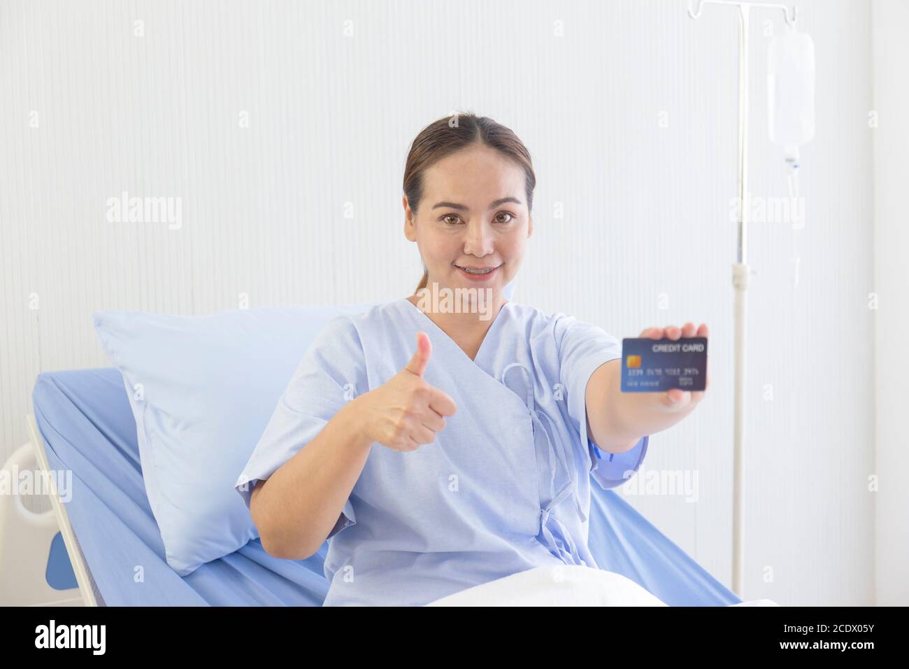 An Asian female patient lay on the hospital bed in her hand, held a credit card, and thumbs up to show a smile. Stock Photo