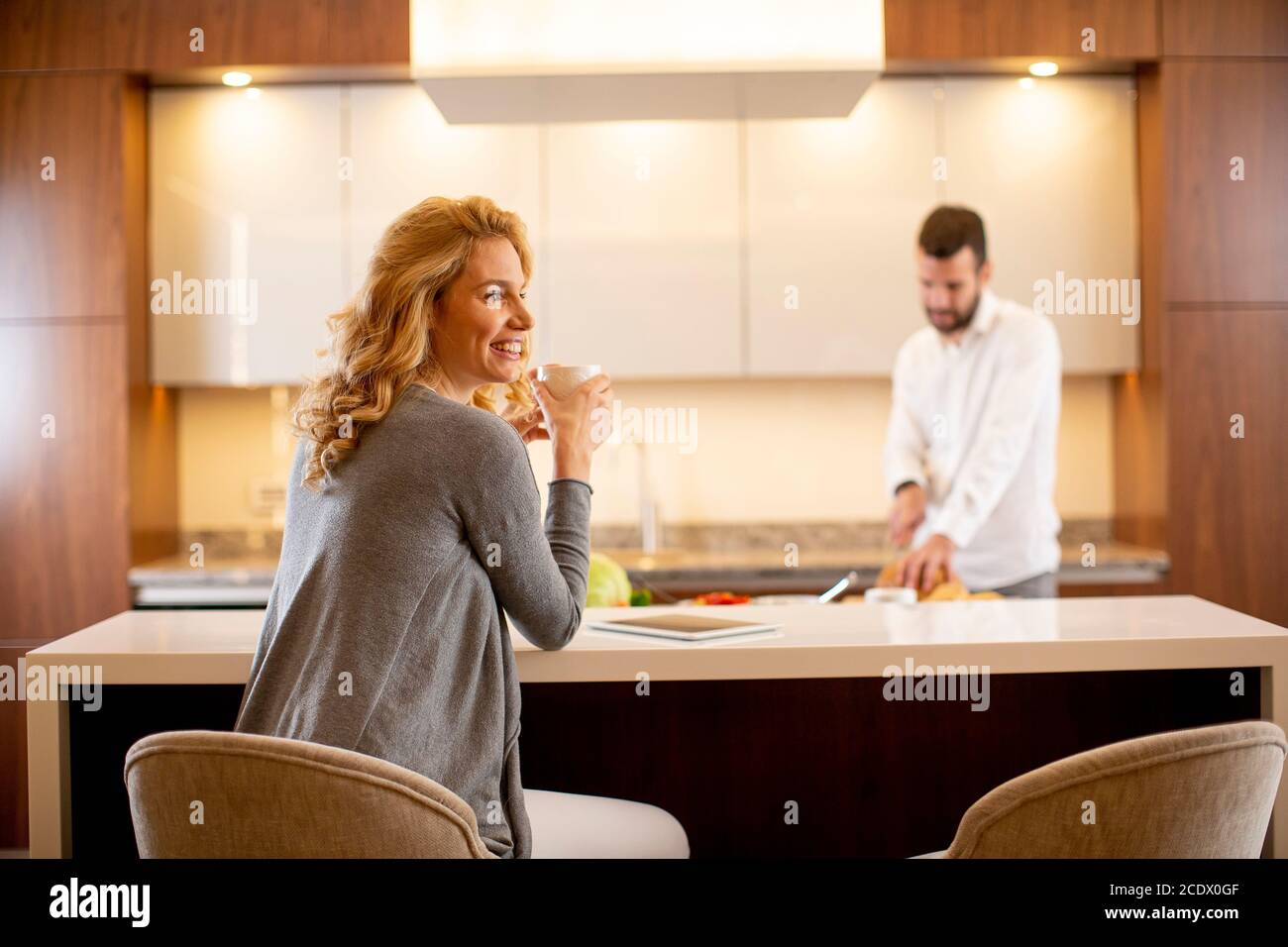 Young woman drinking coffee at the kitchen table while man preparing food Stock Photo