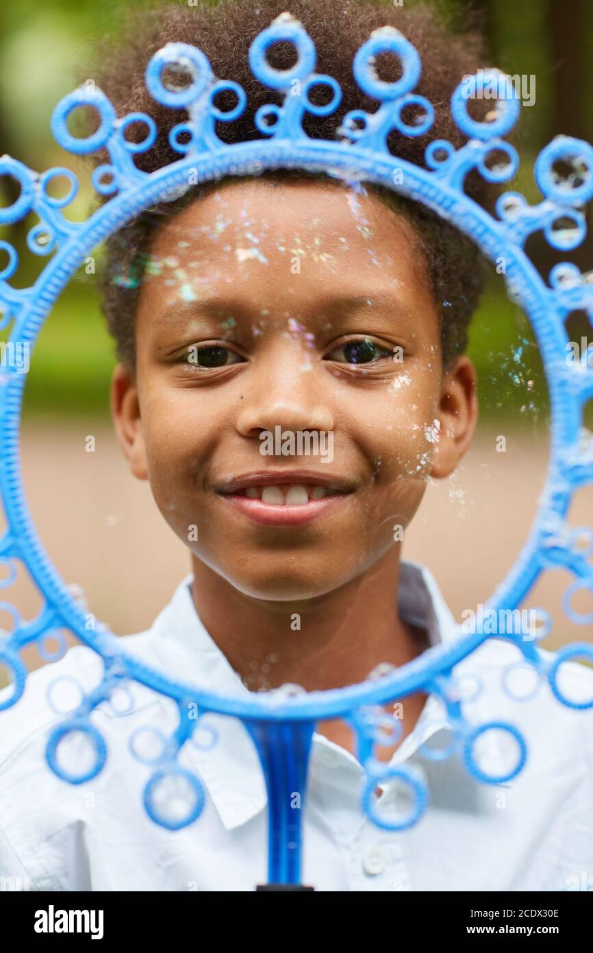 Vertical close up portrait of smiling African-American boy looking at camera through big bubble wand while playing outdoors in park Stock Photo