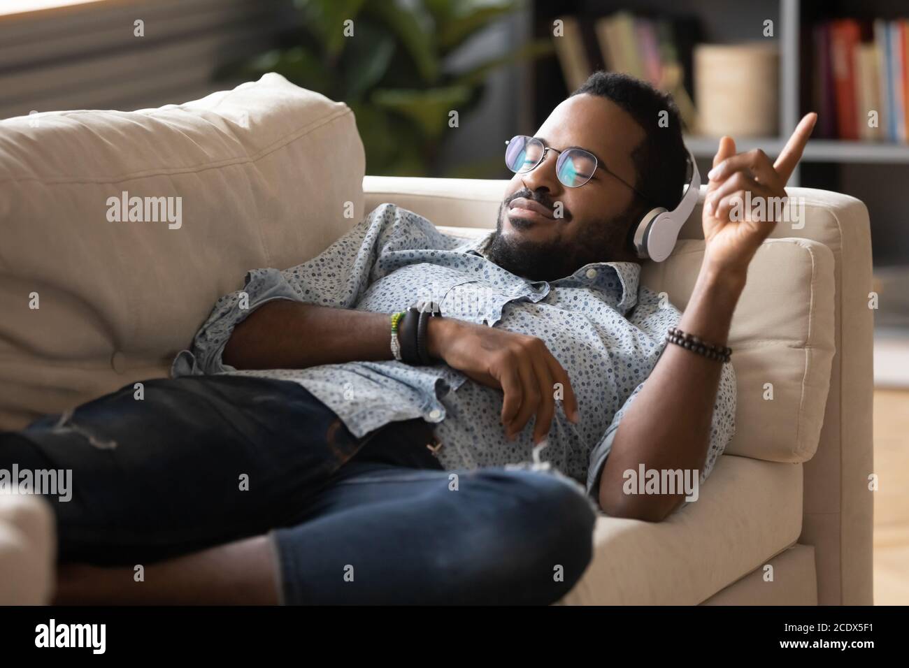 Satisfied positive African American man lying on couch, enjoying music Stock Photo
