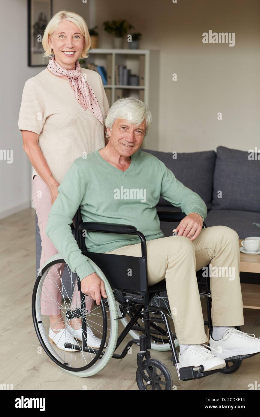 Portrait of senior woman smiling at camera while standing near her disabled husband in wheelchair Stock Photo