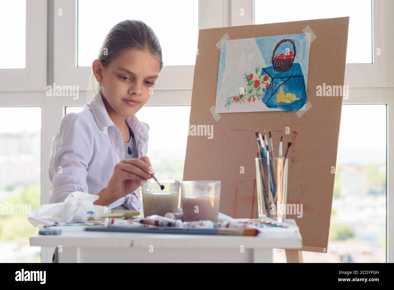 girl sits behind an easel and washes her brush in a glass of water Stock Photo