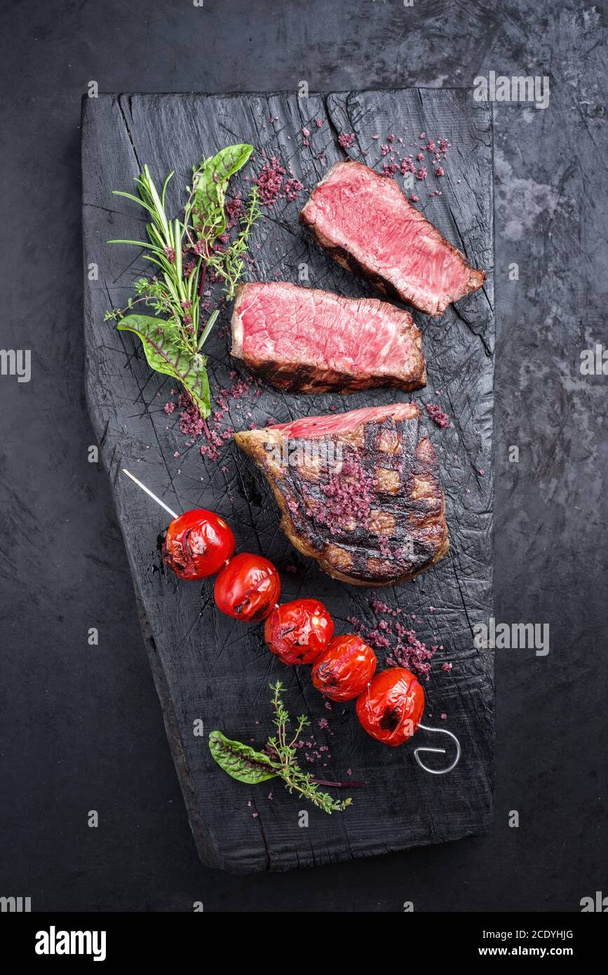 Barbecue dry aged wagyu roast beef steak with tomatoes and herbs as top view on a rustic charred wooden board Stock Photo