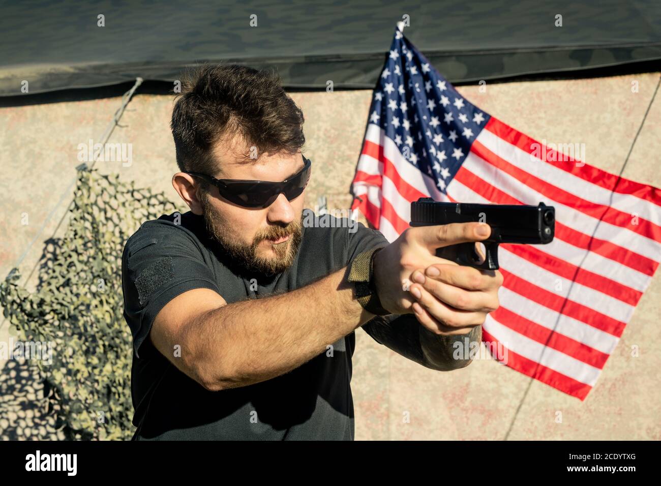 Concentrated American bearded man in sunglasses holding gun and