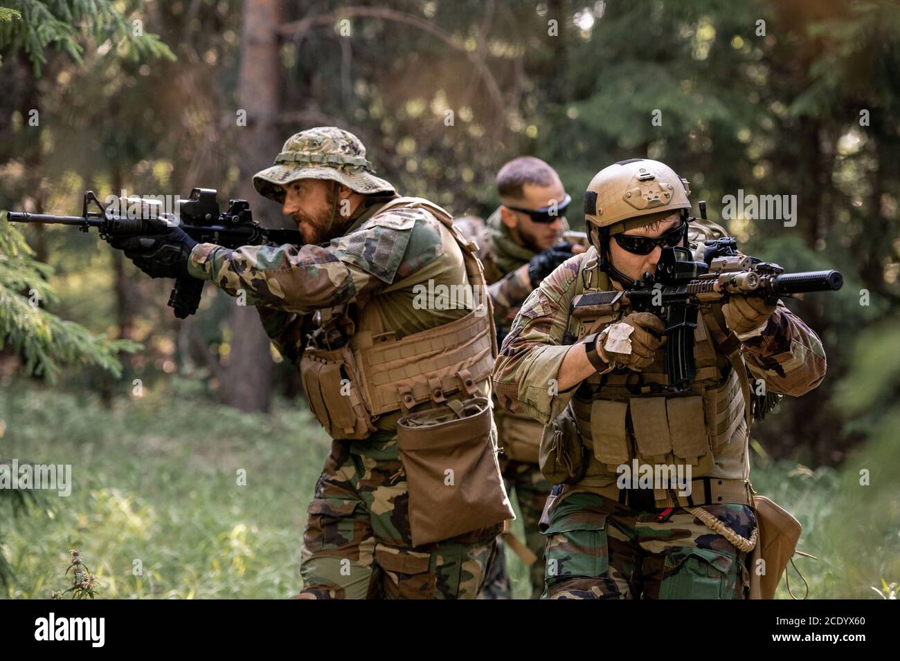 Group of attentive armed soldiers in camouflage outfits moving with rifles in forest while working at clean-up operation Stock Photo