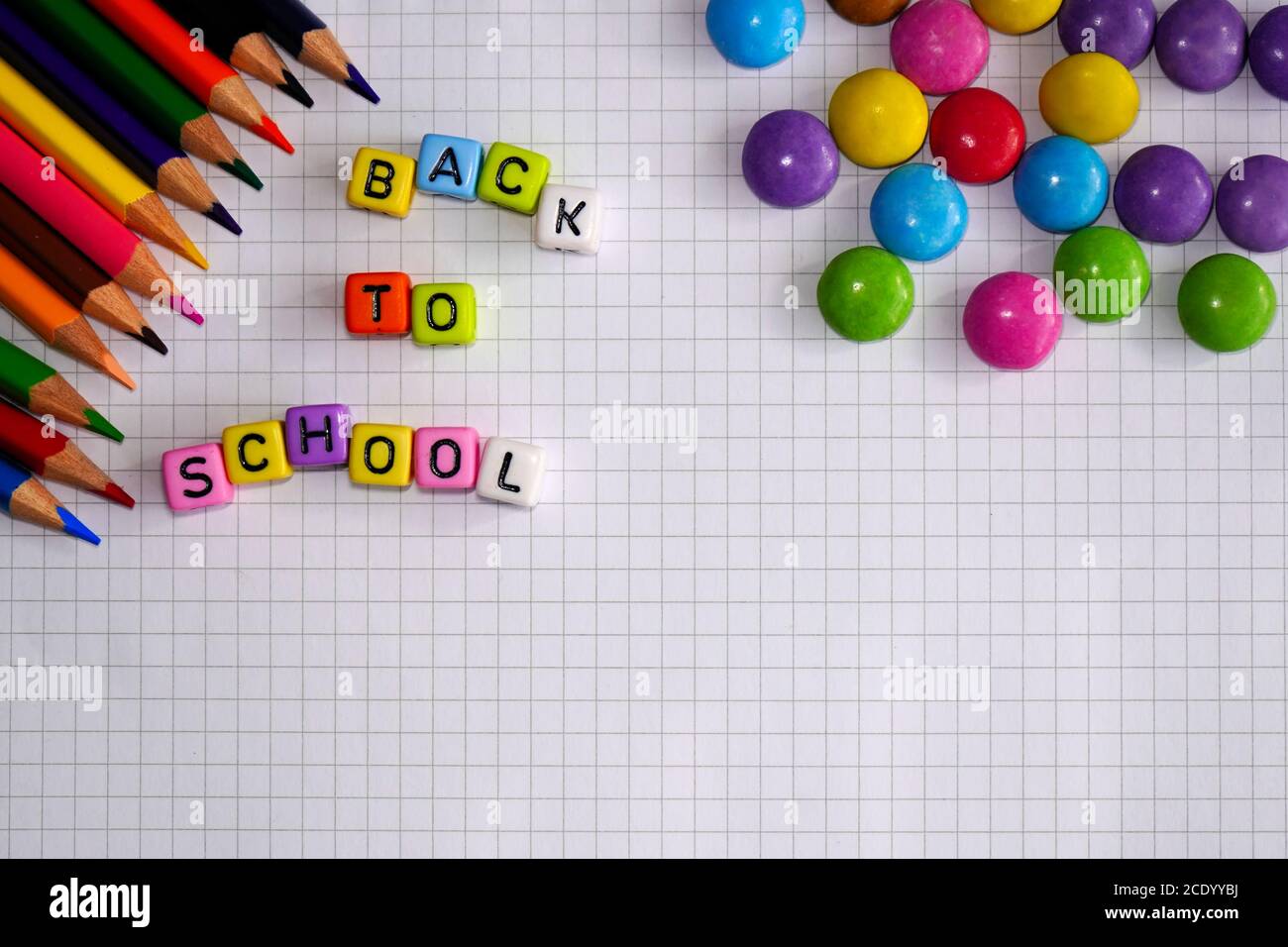 Cheerful 'Back to school' concept with colourful lettering on checked paper. Pencils and chocolate lentils for decoration. Stock Photo