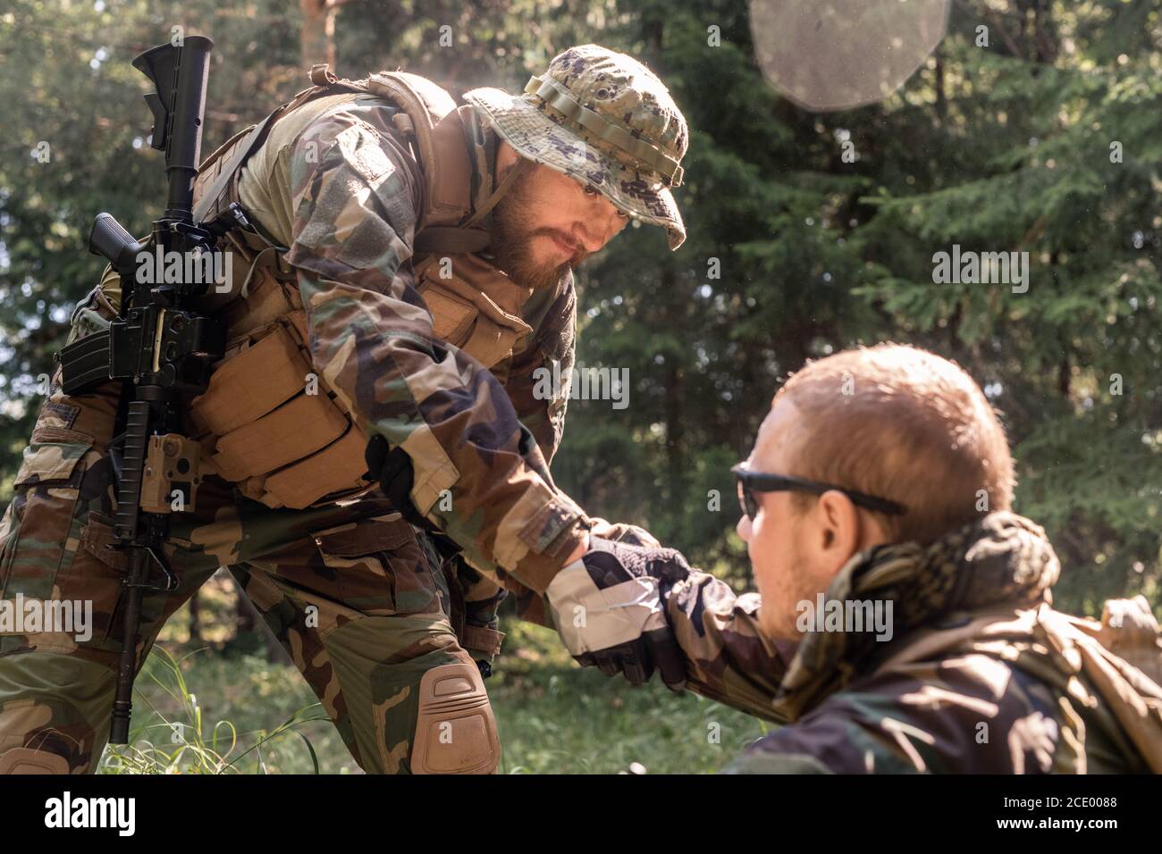 Bearded soldier in hat and vest giving hand to wounded military friend while picking him up off ground in forest Stock Photo