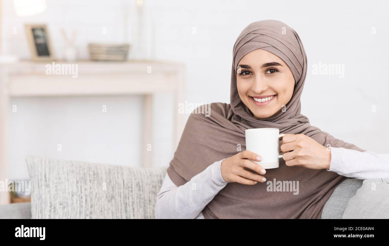 Domestic Relax. Smiling Arabic Girl In Hijab Relaxing On Couch With Coffee Stock Photo