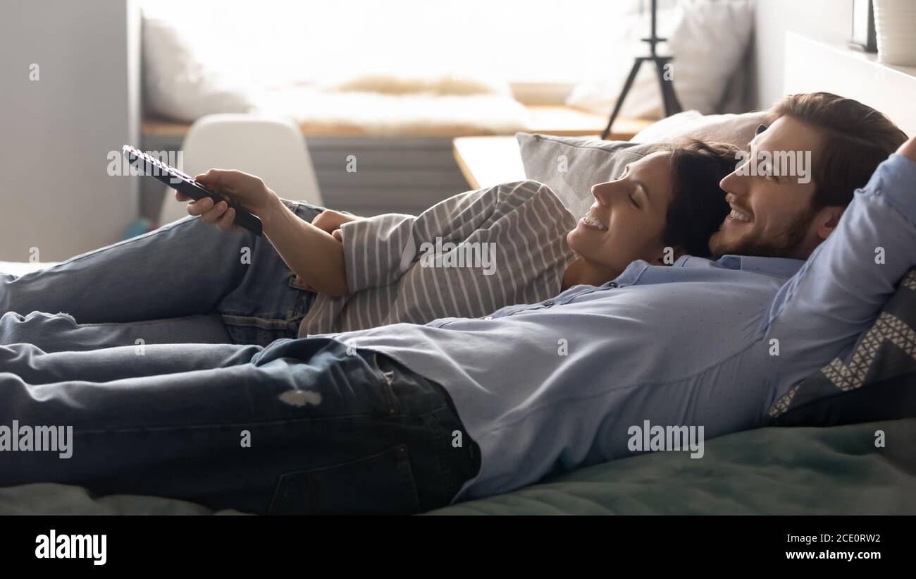 Happy relaxed couple lying on bed, using remote tv controller. Stock Photo