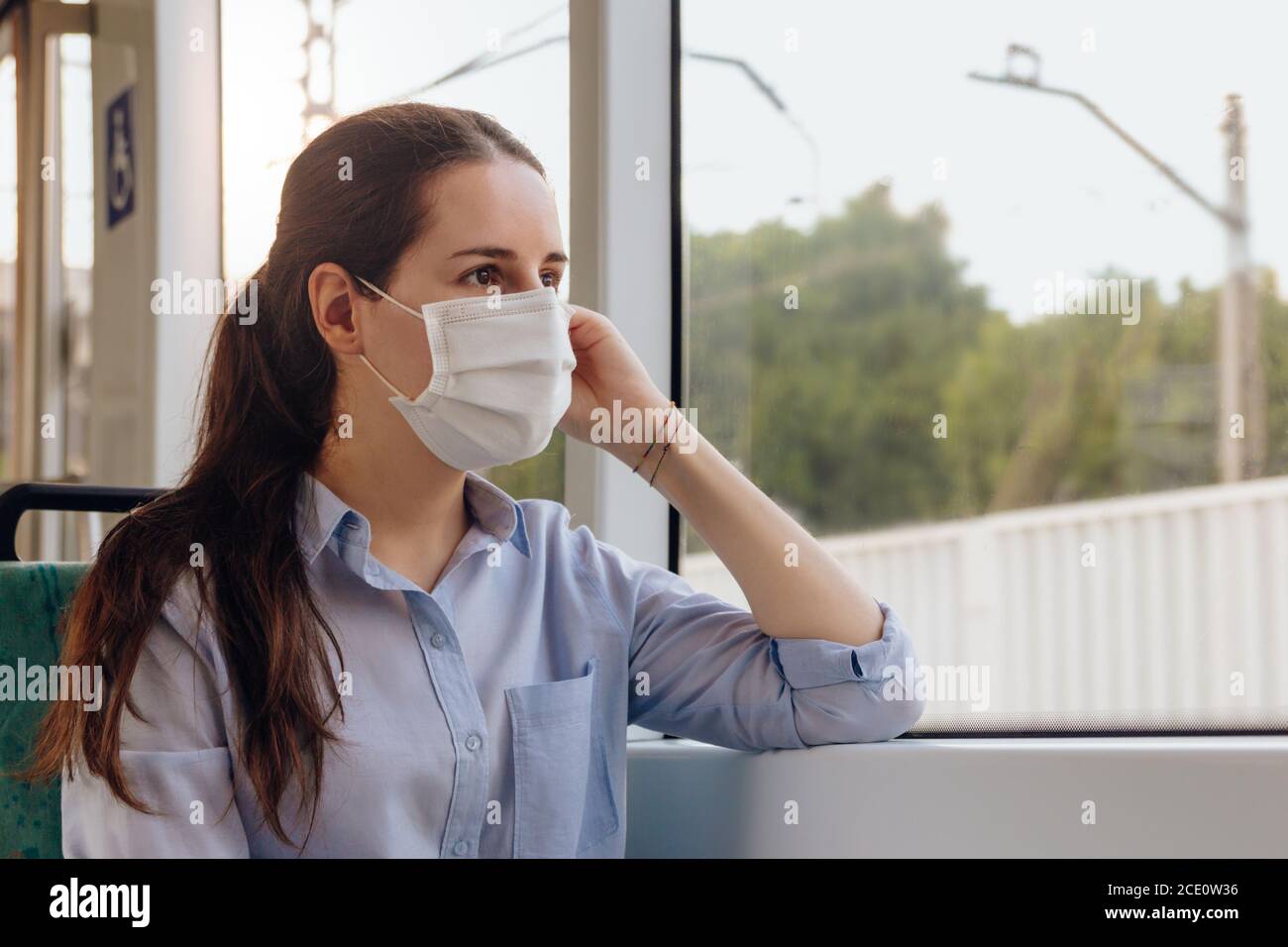 Stock photo of a young woman wearing a face mask traveling by tram. She is looking out the window. New normal concept Stock Photo
