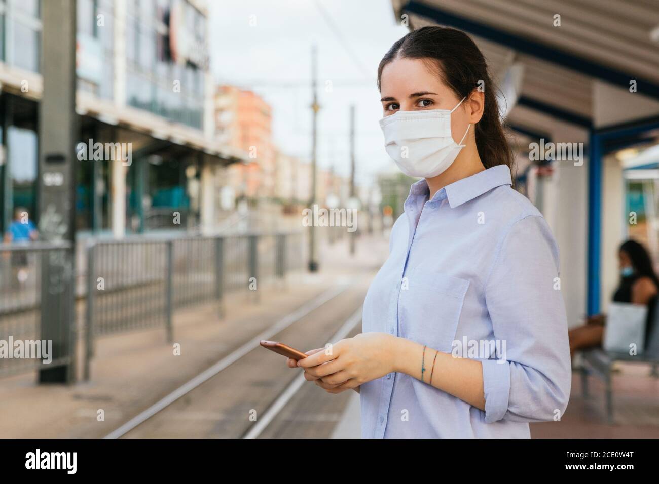 Stock photo of a young woman wearing a face mask, holding a phone and waiting on a tram station. New normal concept Stock Photo