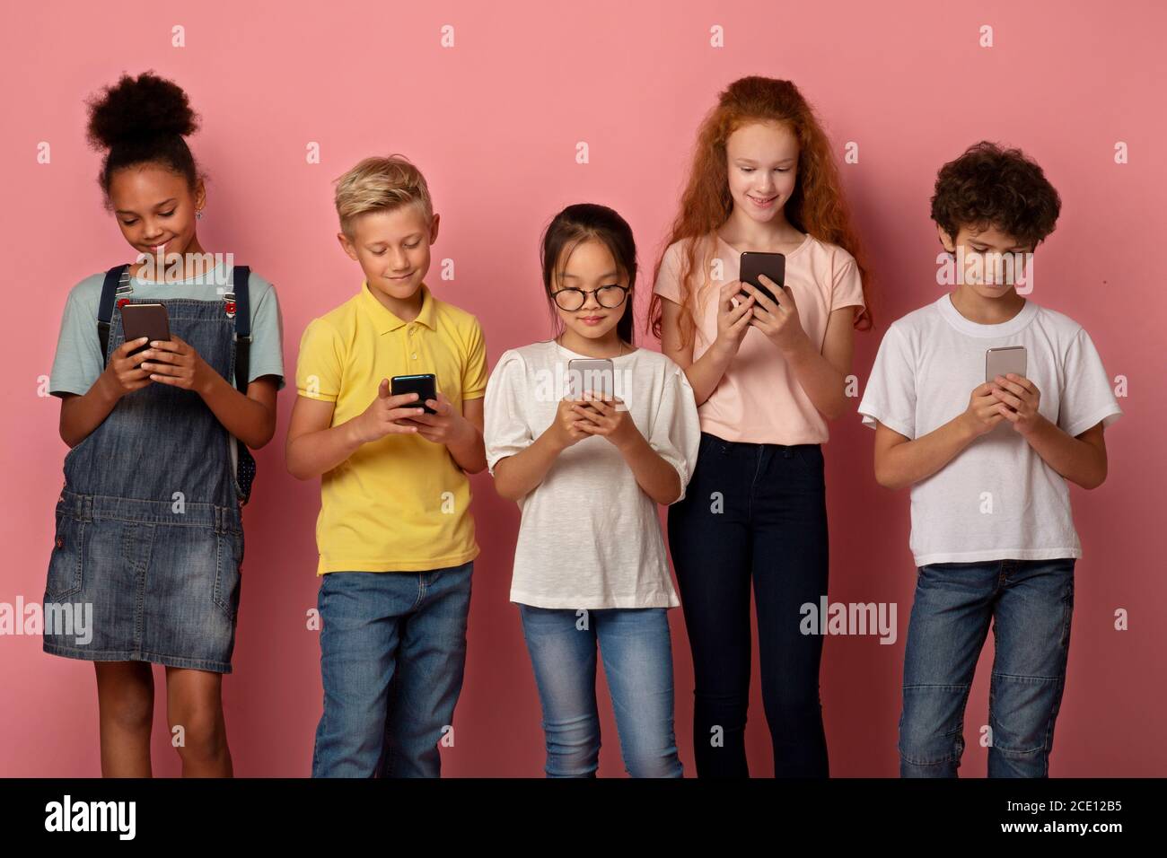 Group of diverse schoolkids stuck in mobile phones studying online on pink background Stock Photo