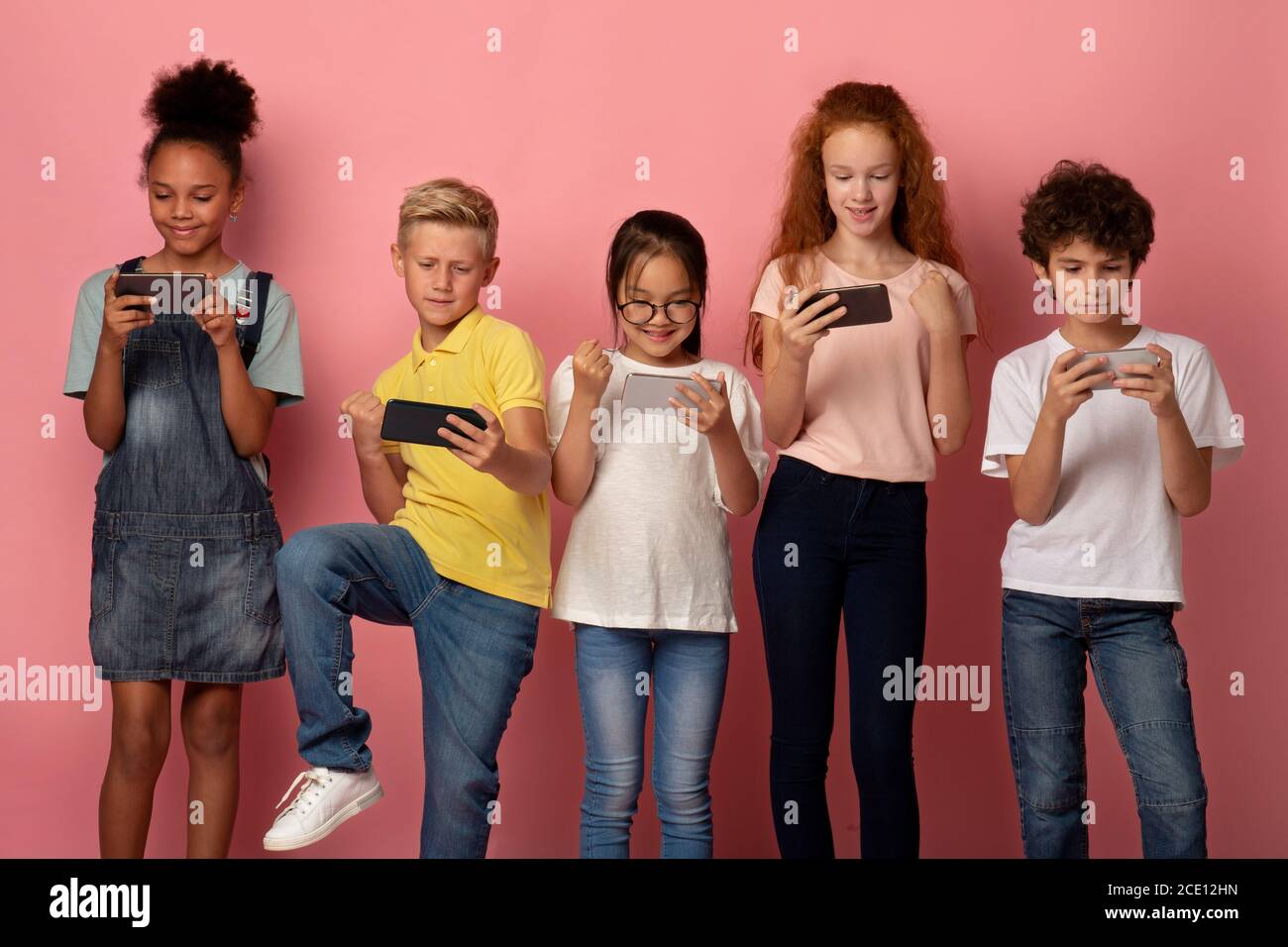 Cheerful schoolchildren playing mobile games on their phones over pink background Stock Photo