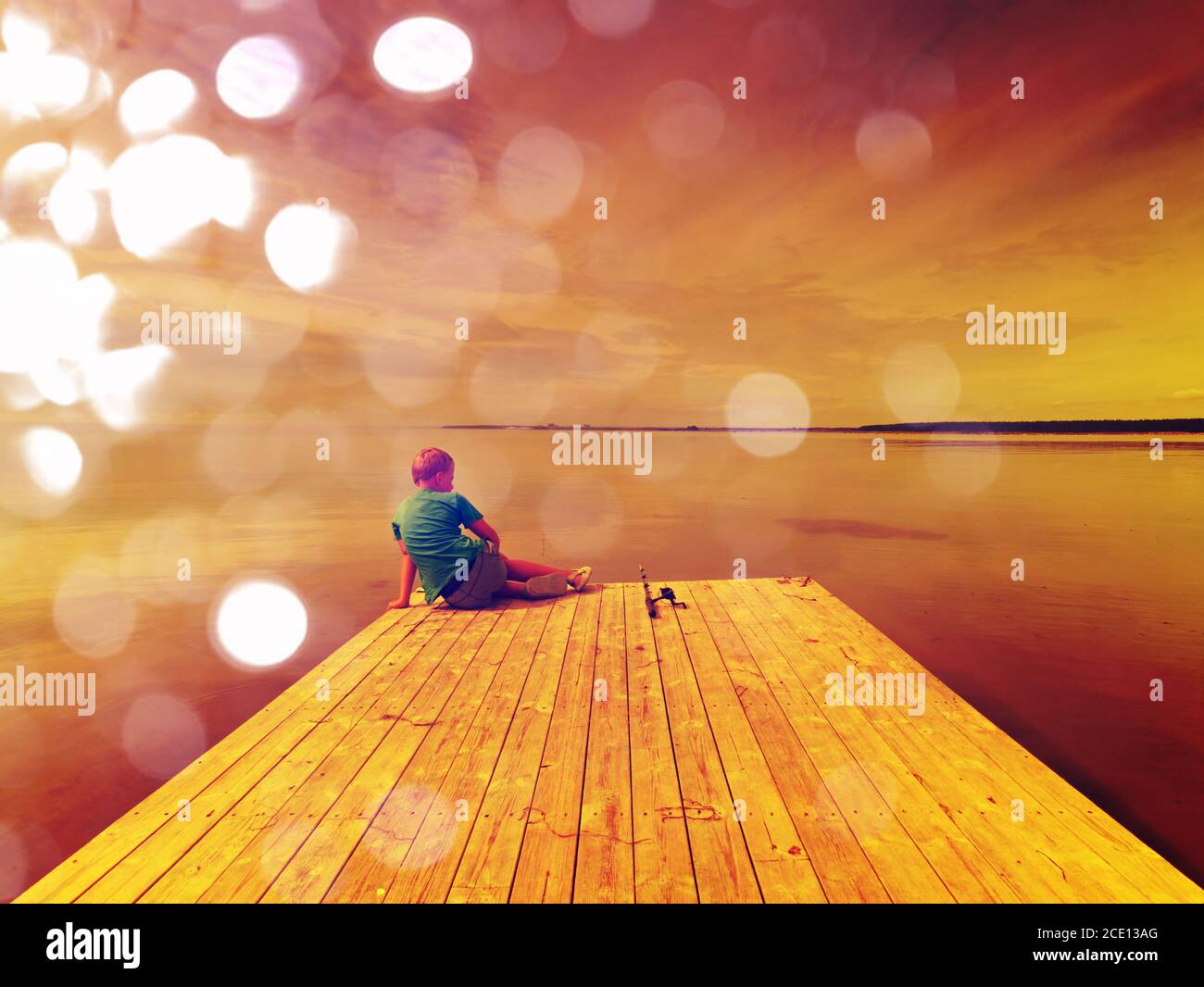 Abstract effect. Small blond hair boy is fishing at the end of wooden mole. Smooth water level in bay Stock Photo