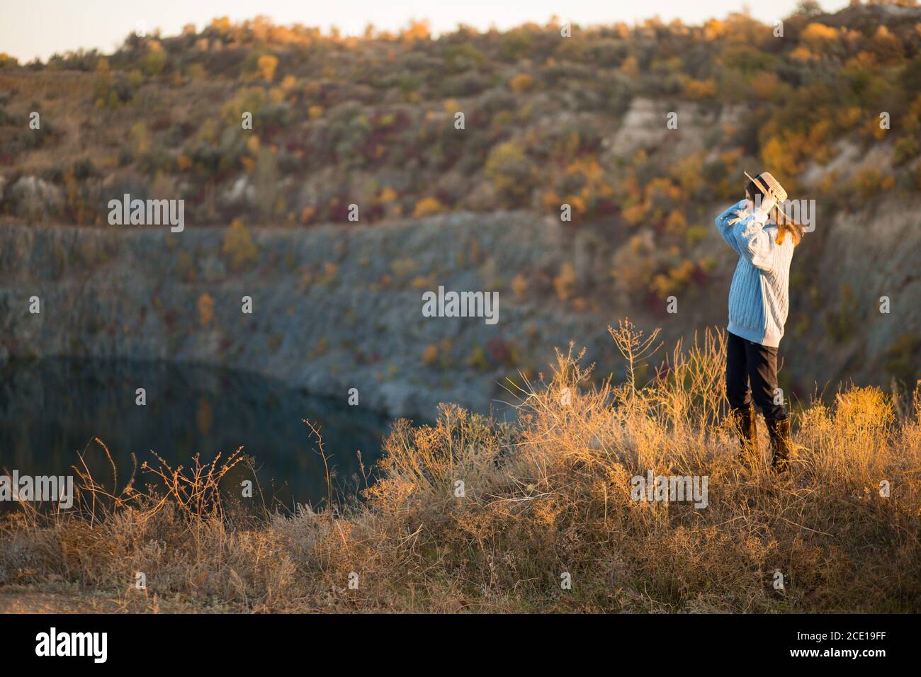Autumn girl in blue sweater and hat standing backwards and admire nature lake view. Autumn forest colors with girl back view. Autumn portrait. Stock Photo